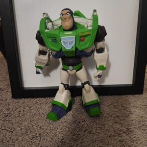 May include: A Buzz Lightyear action figure, featuring a green and white spacesuit with blue and black accents. The figure has a determined expression and is posed in a standing position. The toy is set against a neutral background.