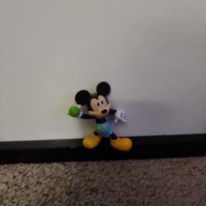 Op de afbeelding: Een Mickey Mouse-beeldje in een dynamische pose, met een groene bal. Het figuur heeft zwarte oren, een rode mond en draagt een blauwe korte broek, gele schoenen en witte handschoenen. De achtergrond is een effen wit oppervlak.