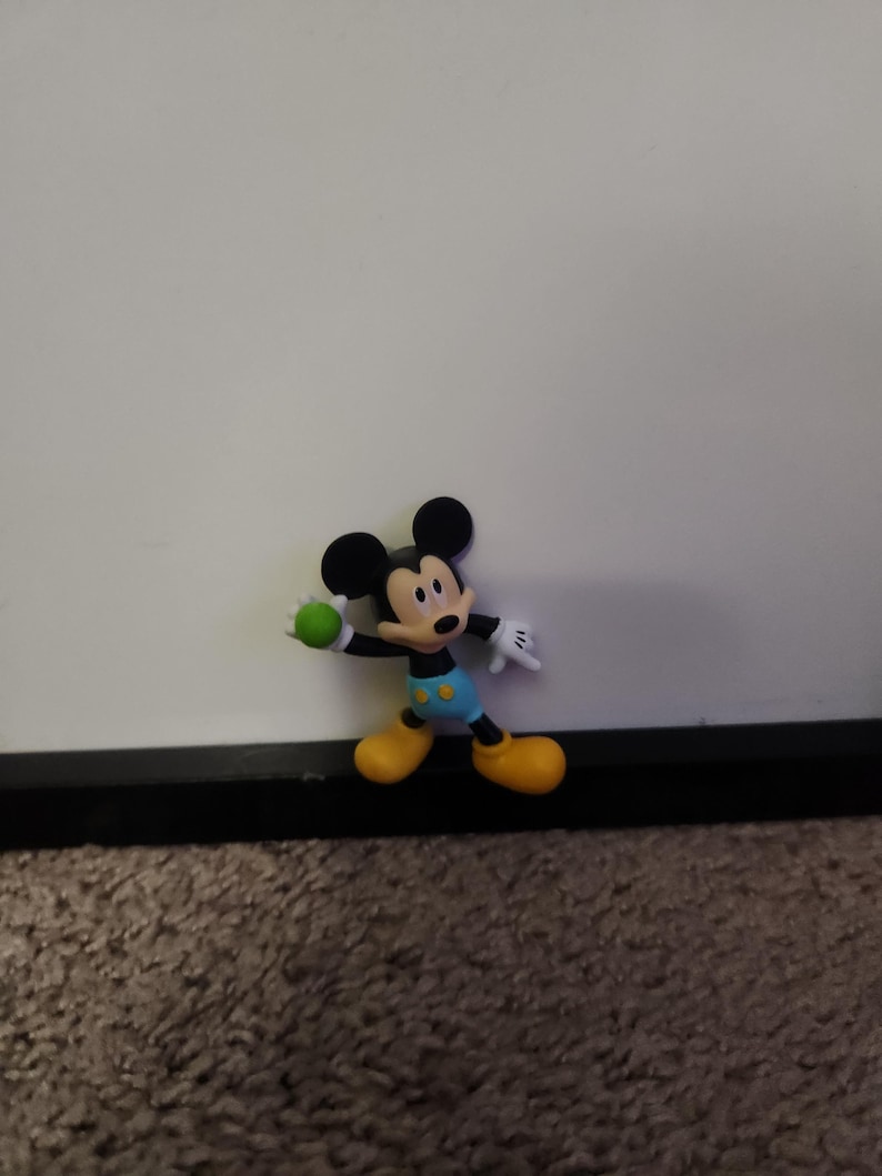 Op de afbeelding: Een Mickey Mouse-beeldje in een dynamische pose, met een groene bal. Het figuur heeft zwarte oren, een rode mond en draagt een blauwe korte broek, gele schoenen en witte handschoenen. De achtergrond is een effen wit oppervlak.