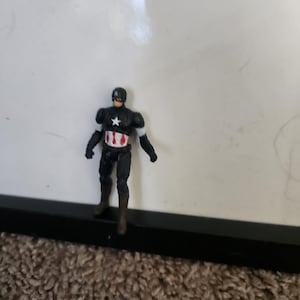 Pu&ograve; includere: Una piccola action figure di Captain America. La figura &egrave; per lo pi&ugrave; nera con accenti bianchi sulle braccia e una stella bianca sul petto. La figura ha un elmetto nero e stivali marroni. La figura &egrave; su una superficie marrone.