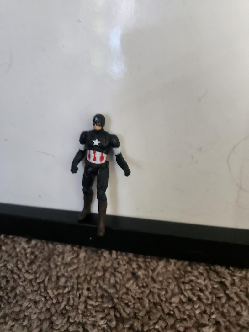 Pu&ograve; includere: Una piccola action figure di Captain America. La figura &egrave; per lo pi&ugrave; nera con accenti bianchi sulle braccia e una stella bianca sul petto. La figura ha un elmetto nero e stivali marroni. La figura &egrave; su una superficie marrone.