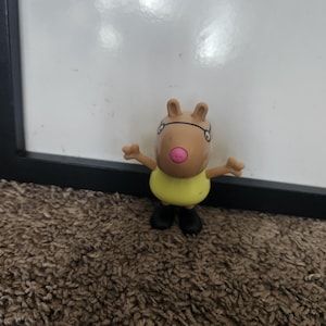 Puede incluir: Pequeña figura de juguete de color marrón claro de un personaje de dibujos animados con una nariz rosa, gafas, una camisa amarilla y zapatos negros. La figura tiene los brazos extendidos. El fondo es una pizarra blanca y una alfombra marrón.