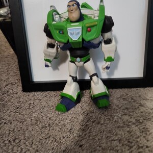 May include: A Buzz Lightyear action figure, featuring a green and white spacesuit with black accents. The figure has a determined expression and is standing in front of a white background.