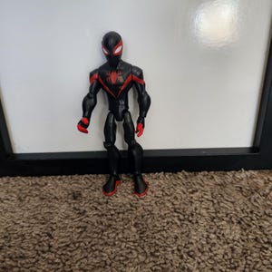 May include: A black and red superhero action figure with a spider emblem. The figure has a black mask with white eyes and red detailing on the gloves and boots. The figure is standing on a brown carpet.