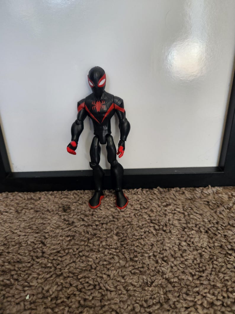 May include: A black and red superhero action figure with a spider emblem. The figure has a black mask with white eye details and red accents on the gloves and boots. The figure is standing on a brown carpet.