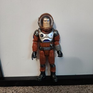 K&ouml;nnte beinhalten: Eine Spielzeug-Astronautenfigur in einem rot-silbernen Raumanzug mit einem durchsichtigen Helm. Die Figur hat schwarze Handschuhe und Stiefel sowie einen silbernen G&uuml;rtel. Der Brustbereich hat ein blau-wei&szlig;es Design.