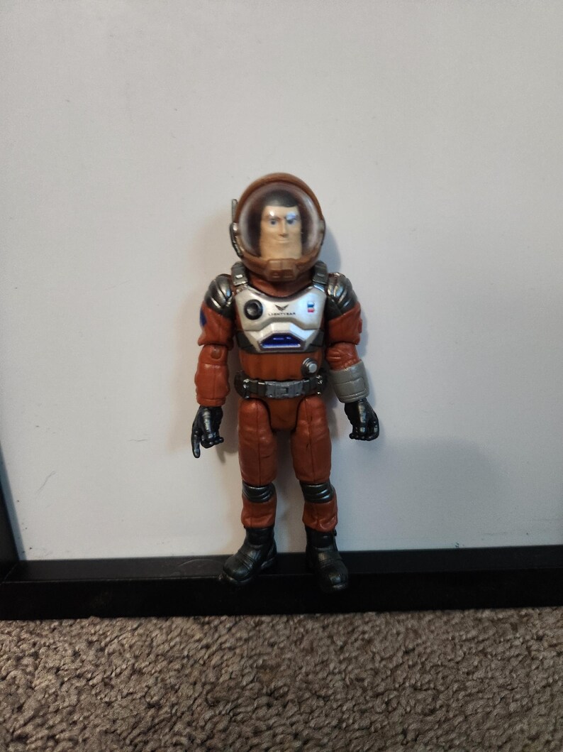 K&ouml;nnte beinhalten: Eine Spielzeug-Astronautenfigur in einem rot-silbernen Raumanzug mit einem durchsichtigen Helm. Die Figur hat schwarze Handschuhe und Stiefel sowie einen silbernen G&uuml;rtel. Der Brustbereich hat ein blau-wei&szlig;es Design.