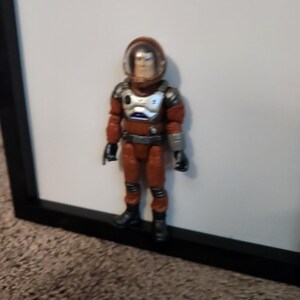 K&ouml;nnte beinhalten: Vintage Actionfigur eines Astronauten in einem rot-silbernen Raumanzug mit transparentem Helm. Die Figur steht vor einem wei&szlig;en Hintergrund und einem schwarzen Rahmen. Sie hat schwarze Stiefel und Handschuhe.