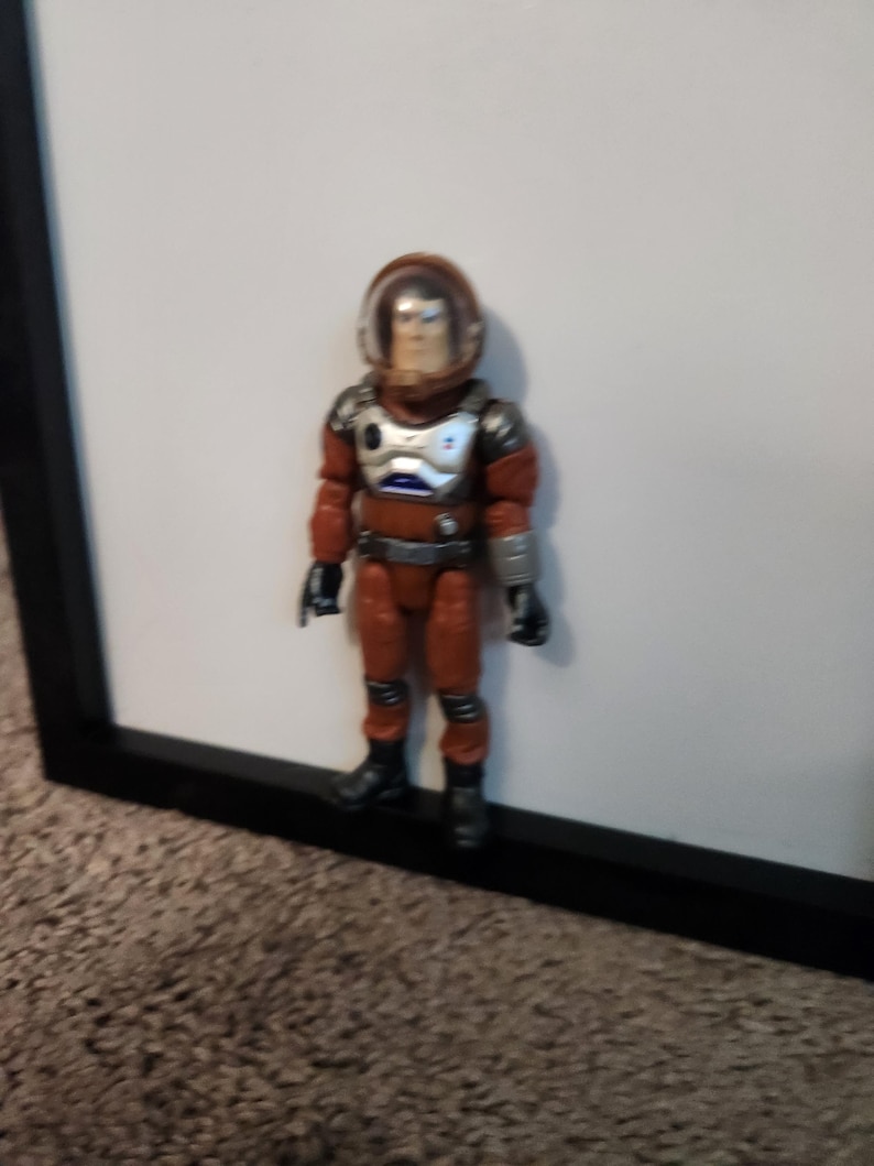 K&ouml;nnte beinhalten: Vintage Actionfigur eines Astronauten in einem rot-silbernen Raumanzug mit transparentem Helm. Die Figur steht vor einem wei&szlig;en Hintergrund und einem schwarzen Rahmen. Sie hat schwarze Stiefel und Handschuhe.