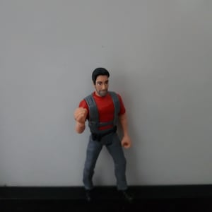 Può includere: Piccola action figure di un uomo con una maglietta rossa, una tuta grigia e stivali neri. La figura ha una barba scura e capelli corti neri. La figura è in posa con un pugno chiuso.