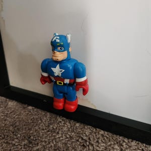 May include: A Captain America action figure in a stylized design. The figure is primarily blue with red gloves and boots. A white star is on the chest. The figure is standing in front of a white background.