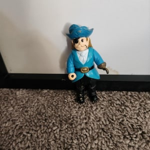 May include: A small plastic pirate figurine. The pirate wears a blue coat, a white shirt, and black pants. He has a blue hat, an eye patch, and a hook for a hand. The figurine is standing on a brown carpet.