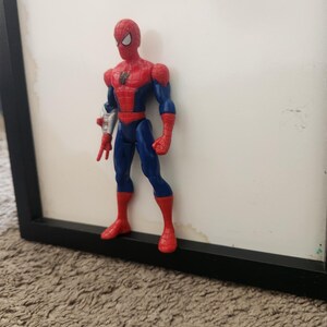 Może przedstawiać: Figurka akcji Spider-Mana w czerwono-niebieskim kostiumie z czarnym emblematem pająka na klatce piersiowej. Figurka posiada srebrny dodatek na prawym przedramieniu, białe soczewki oczu i stoi w wyprostowanej pozycji w czerwonych butach.