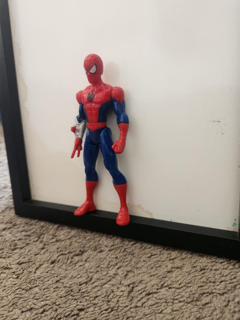 Może przedstawiać: Figurka akcji Spider-Mana w czerwono-niebieskim kostiumie z czarnym emblematem pająka na klatce piersiowej. Figurka posiada srebrny dodatek na prawym przedramieniu, białe soczewki oczu i stoi w wyprostowanej pozycji w czerwonych butach.