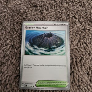 Puede incluir: Una carta de Entrenador Gravity Mountain del juego de cartas Pokémon. La carta presenta una ilustración de una montaña rodeada de agua. El texto de la carta incluye instrucciones del juego y el nombre del artista.