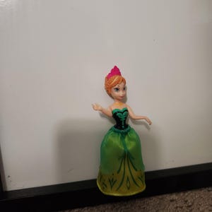 May include: A small princess doll in a green and gold dress with a black bodice. The doll has orange hair and a pink crown. The doll is positioned against a white backdrop.