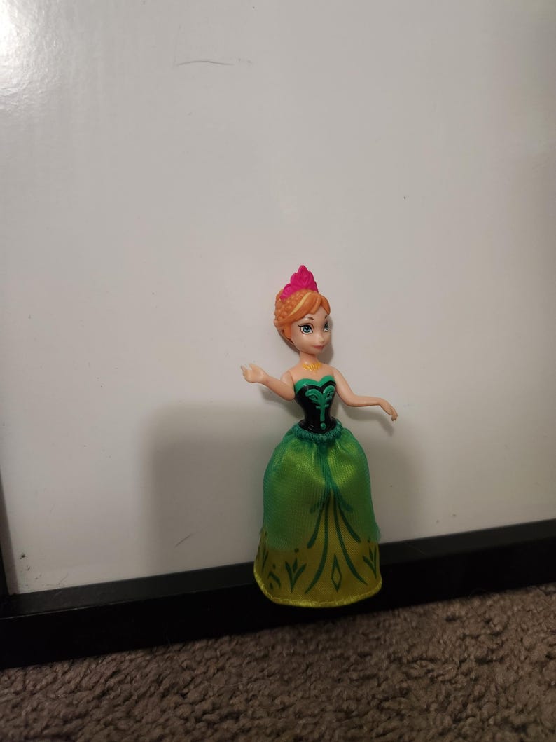 May include: A small princess doll in a green and gold dress with a black bodice. The doll has orange hair and a pink crown. The doll is positioned against a white backdrop.