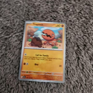 Może przedstawiać: Karta kolekcjonerska Pokémon z Trapinchem, pomarańczowym stworzeniem z pękniętą głową, na tle pustynnego krajobrazu. Karta zawiera tekst "Call for Family" i "Bite", z wartością obrażeń 20.