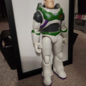 May include: A Buzz Lightyear action figure, featuring a white and green spacesuit with purple accents. The figure has a detailed face and is standing upright. The toy is a collectible item.