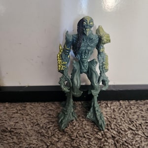 Peut inclure: Figurine d'action détaillée d'une créature humanoïde avec un corps gris-vert, des accents dorés et une longue tresse sombre. La figurine a une apparence squelettique et est posée avec les bras le long du corps. La figurine mesure environ 15 cm de haut.