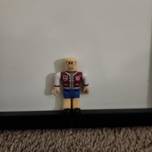 May include: A small plastic figurine of a person wearing a red and white letterman jacket with blue jeans and a white shirt. The figurine has a smiling face with glasses and braces.