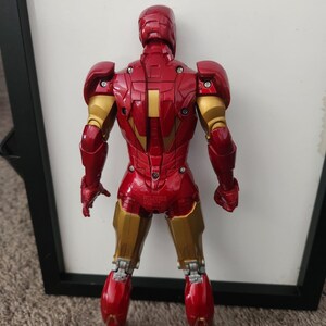 May include: A red and gold Iron Man action figure shown from the back, featuring articulated joints, visible screws on the armor plates, and a metallic finish. The figure stands upright with arms at its sides, displaying detailed mechanical plating.