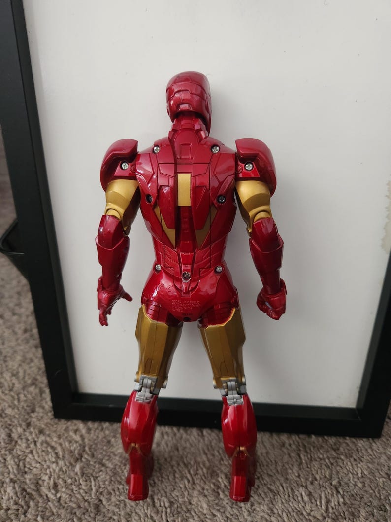 May include: A red and gold Iron Man action figure shown from the back, featuring articulated joints, visible screws on the armor plates, and a metallic finish. The figure stands upright with arms at its sides, displaying detailed mechanical plating.