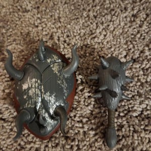 May include: A gray and red shield with curved horns and a matching mace with spikes. The shield has a distressed metallic finish. The mace has a wooden handle and a spiked head. The items are likely toy props.