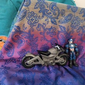 May include: A silver toy motorcycle and a Captain America action figure rest on a paisley patterned fabric with blue, purple, and pink hues. A teal fabric is visible in the background.
