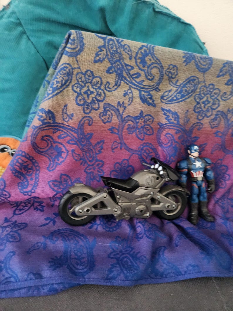 May include: A silver toy motorcycle and a Captain America action figure rest on a paisley patterned fabric with blue, purple, and pink hues. A teal fabric is visible in the background.