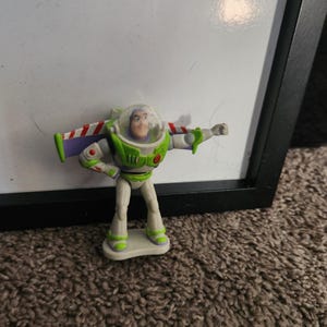 Puede incluir: Una peque&ntilde;a figura de acci&oacute;n de Buzz Lightyear. La figura es principalmente blanca y verde, con alas a rayas rojas y blancas. Tiene un casco de pl&aacute;stico transparente y est&aacute; posando con un brazo extendido. La figura est&aacute; sobre una peque&ntilde;a base blanca.