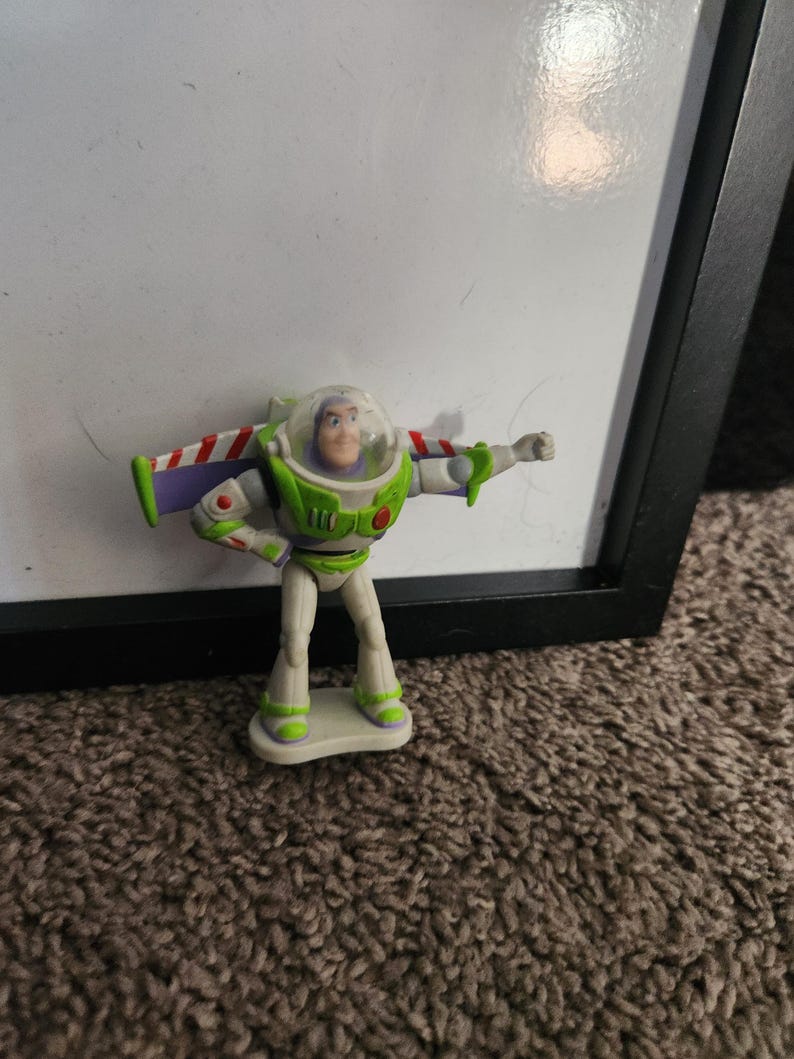 Puede incluir: Una peque&ntilde;a figura de acci&oacute;n de Buzz Lightyear. La figura es principalmente blanca y verde, con alas a rayas rojas y blancas. Tiene un casco de pl&aacute;stico transparente y est&aacute; posando con un brazo extendido. La figura est&aacute; sobre una peque&ntilde;a base blanca.
