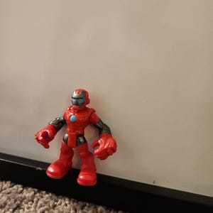 May include: A red and black action figure of a superhero with a helmet, gloves, and boots. The figure has a blue circle on its chest and is standing on a carpeted surface.