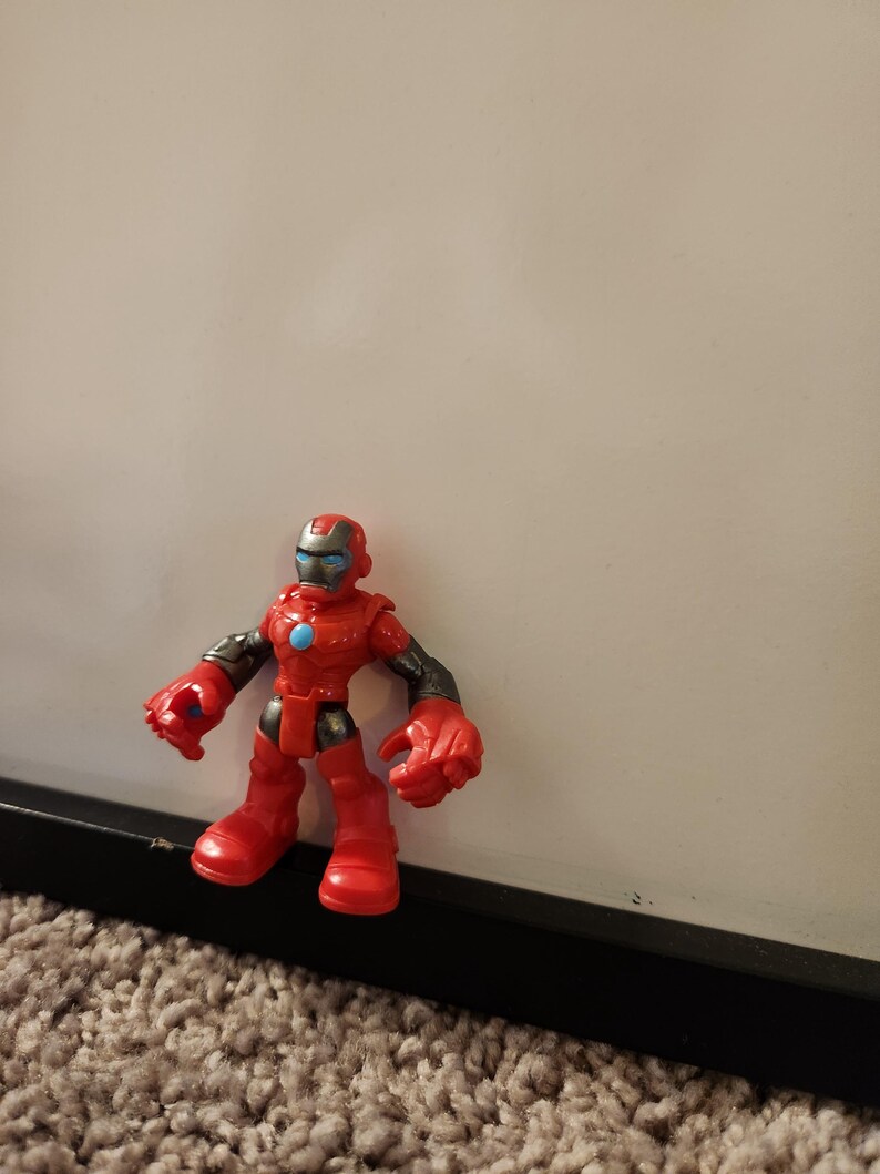 May include: A red and black action figure of a superhero with a helmet, gloves, and boots. The figure has a blue circle on its chest and is standing on a carpeted surface.