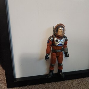 K&ouml;nnte beinhalten: Eine Spielzeug-Astronautenfigur in einem orangefarbenen Raumanzug mit einem transparenten Helm. Die Figur hat schwarze Handschuhe und Stiefel mit silbernen und blauen Akzenten auf Brust und Armen. Die Figur steht vor einem wei&szlig;en Hintergrund.