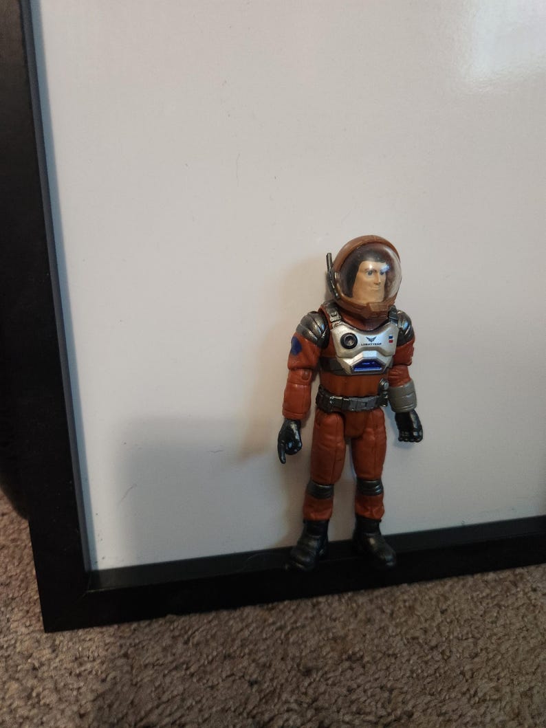 K&ouml;nnte beinhalten: Eine Spielzeug-Astronautenfigur in einem orangefarbenen Raumanzug mit einem transparenten Helm. Die Figur hat schwarze Handschuhe und Stiefel mit silbernen und blauen Akzenten auf Brust und Armen. Die Figur steht vor einem wei&szlig;en Hintergrund.