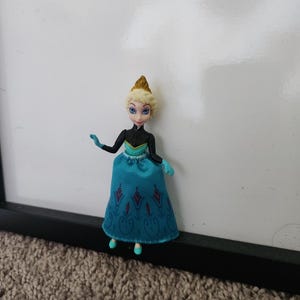 May include: A small plastic doll of a princess wearing a blue and black dress with a crown. The doll has blonde hair and blue eyes.
