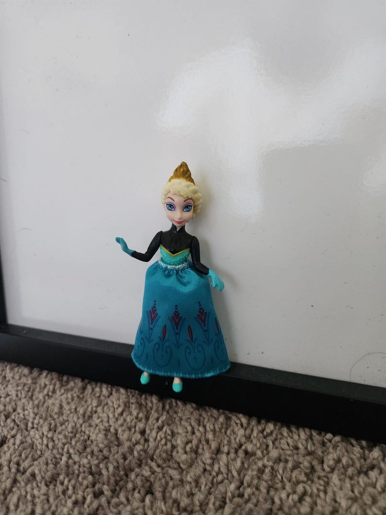 May include: A small plastic doll of a princess wearing a blue and black dress with a crown. The doll has blonde hair and blue eyes.