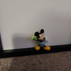 Op de afbeelding: Een Mickey Mouse-beeldje in een staande pose, met een groene bal. Het figuur heeft zwarte oren, een zwart gezicht en een witte handschoen. Mickey draagt gele schoenen, een blauwe korte broek met gele knopen en een zwart-wit shirt.