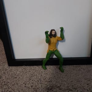 May include: A small action figure of a superhero with long brown hair and a beard. The figure is wearing a yellow and green costume with green gloves and boots. The figure is posed with arms raised.