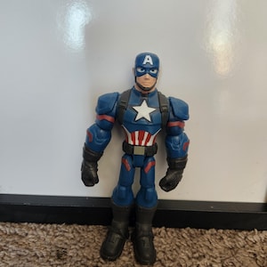 May include: A Captain America action figure. The figure is primarily blue with red and white accents. It features a helmet with a white "A", a white star on the chest, and black gloves and boots. The figure is standing on a brown carpet.