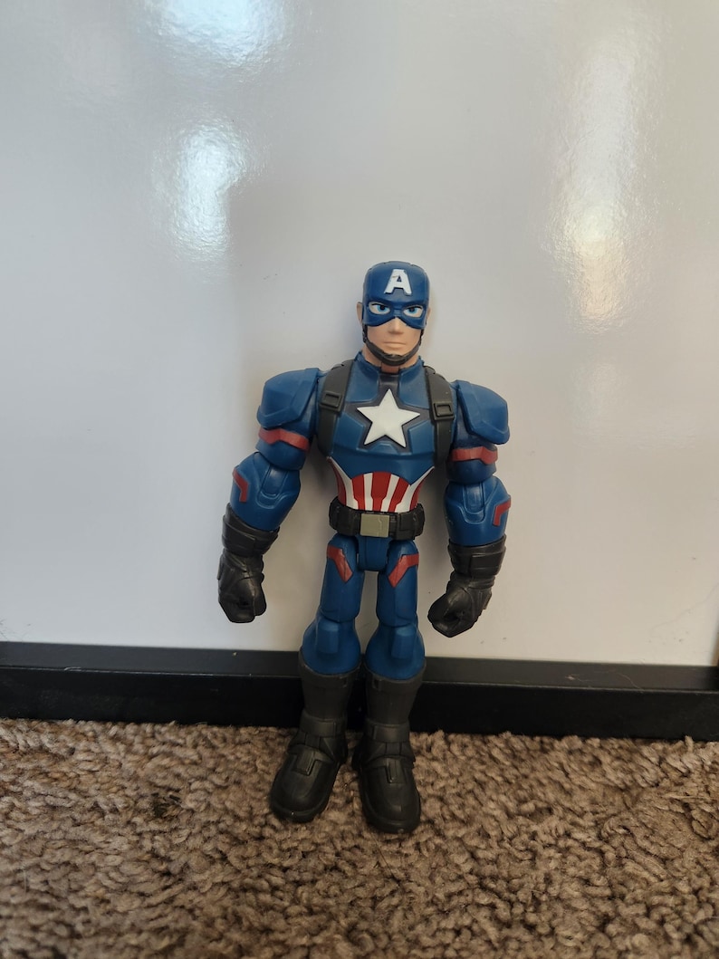 May include: A Captain America action figure. The figure is primarily blue with red and white accents. It features a helmet with a white "A", a white star on the chest, and black gloves and boots. The figure is standing on a brown carpet.