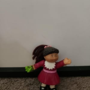 Peut inclure: Une petite figurine en plastique représentant un enfant aux cheveux en laine brun foncé attachés en queue de cheval avec un nœud rose. Le personnage porte une robe bordeaux à manches longues avec un col en dentelle blanche, une ceinture rose, des chaussettes blanches et des chaussures noires, tenant un objet vert.