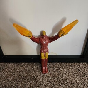 May include: A red and gold Iron Man action figure with outstretched arms and yellow energy blasts. The figure is standing on a brown carpet, with a white background and a black frame.