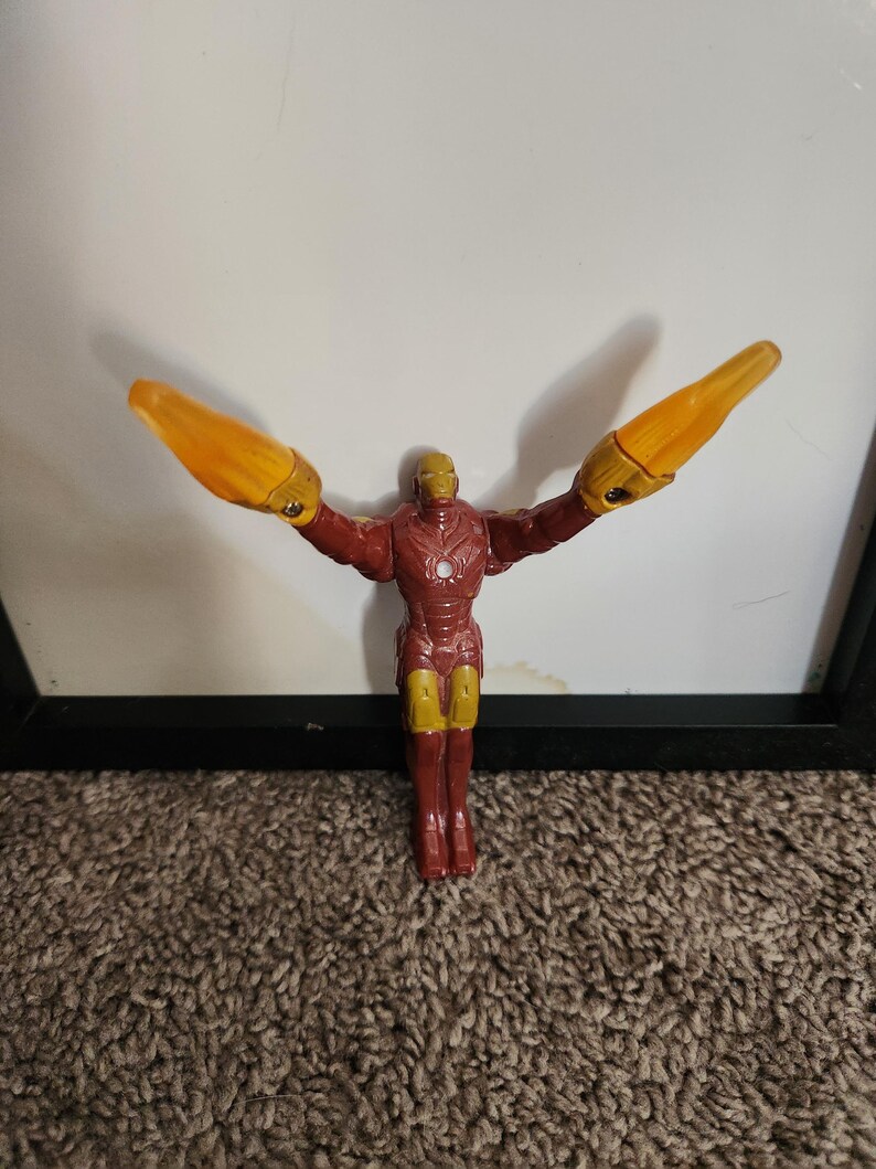 May include: A red and gold Iron Man action figure with outstretched arms and yellow energy blasts. The figure is standing on a brown carpet, with a white background and a black frame.