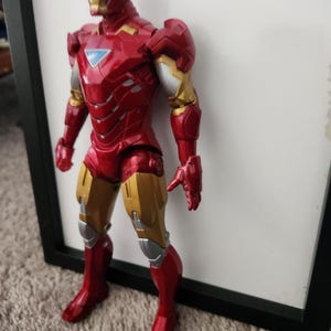 May include: An Iron Man action figure featuring a metallic red and gold suit with silver accents. The character has a triangular light blue chest piece, a full face mask, and articulated limbs, standing in a vertical position with a detailed armor design.