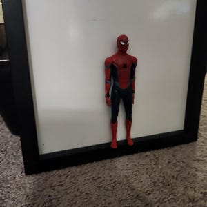 Op de afbeelding: Een Spider-Man actiefiguur in een rood en blauw pak, staand voor een witte achtergrond in een zwart frame. De figuur heeft een gedetailleerd ontwerp, met een masker en een dynamische pose, die de iconische superheld laat zien.