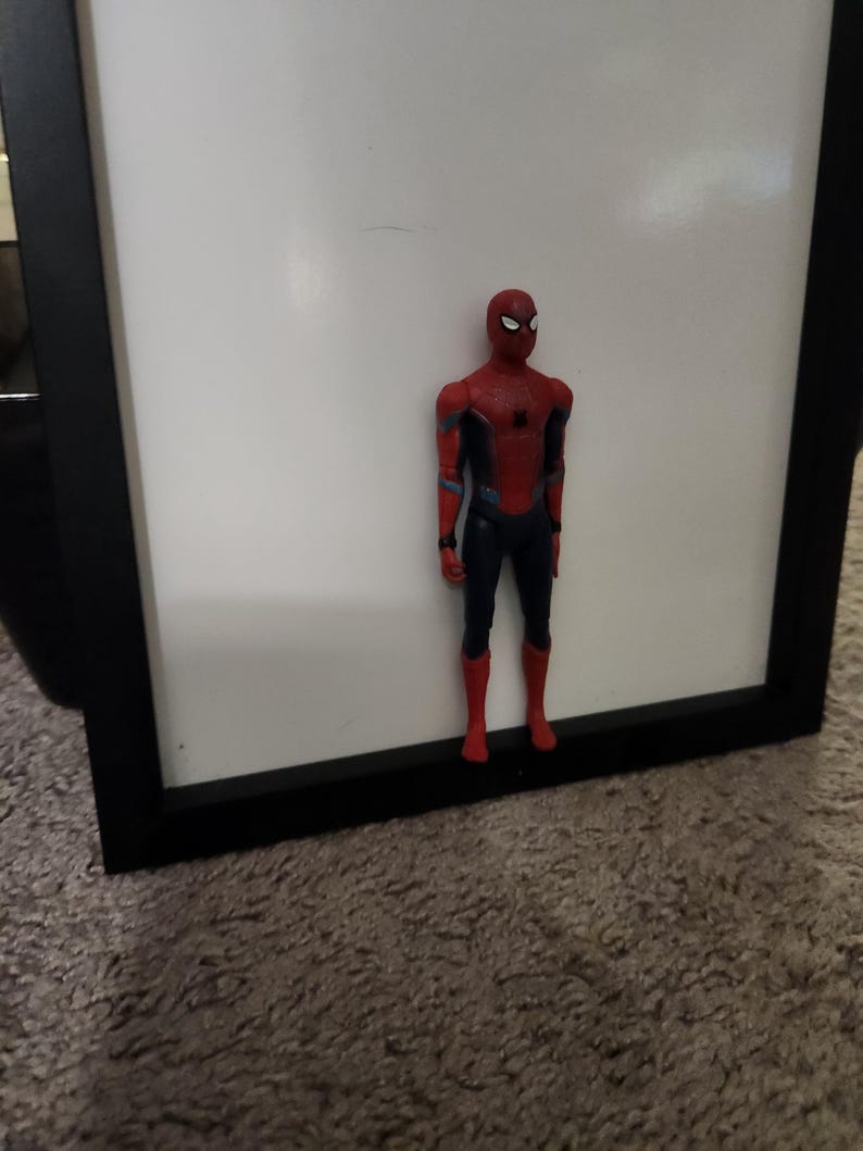 Op de afbeelding: Een Spider-Man actiefiguur in een rood en blauw pak, staand voor een witte achtergrond in een zwart frame. De figuur heeft een gedetailleerd ontwerp, met een masker en een dynamische pose, die de iconische superheld laat zien.