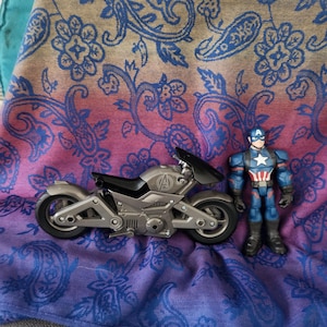 May include: A gray toy motorcycle with black accents and the Avengers logo, next to a Captain America action figure. The items are on a paisley patterned fabric with blue, pink, and tan colors.