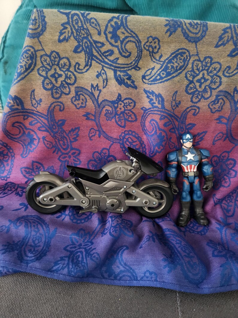 May include: A gray toy motorcycle with black accents and the Avengers logo, next to a Captain America action figure. The items are on a paisley patterned fabric with blue, pink, and tan colors.